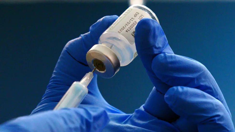 A gloved hand inserts a needle into a vaccination bottle.