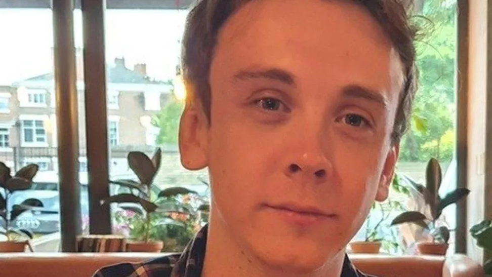 A close-up of Jacob Billington showing his face in front of a row of a plants and a window