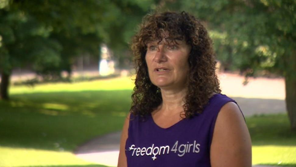 Tina Leslie, standing in a park wearing a purple singlet top, bearing the logo Freedom4Girls. She is standing in a park with trees in the background.