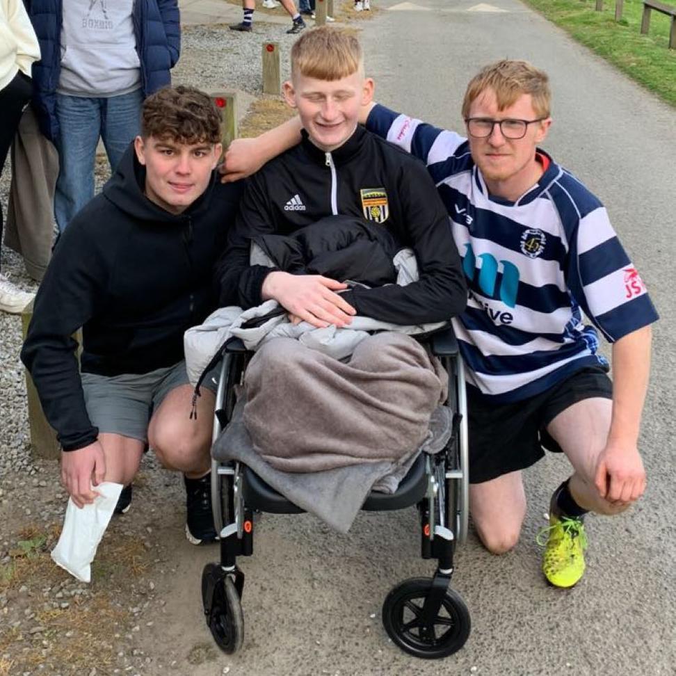 Adam Golebiewski in a wheelchair, smiling, flanked by two young men with their arms round him.