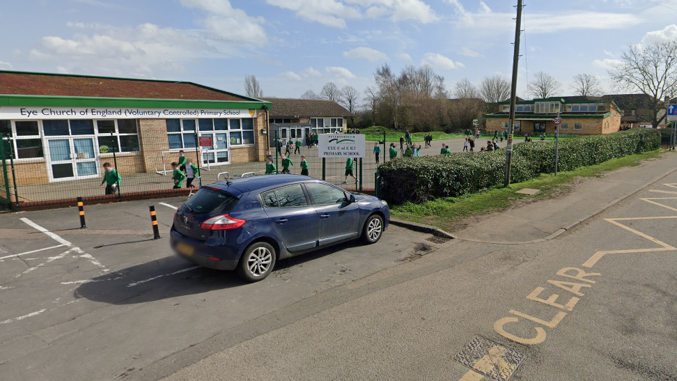 School building with children wearing green jumpers playing outside. A blue car is parked in front of the school.