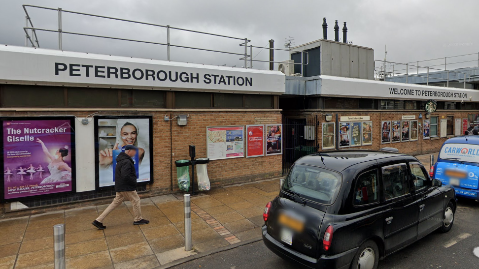 Street view image of Peterborough Train Station. The sky is a dark grey and there are several taxis parked outside the entrance. There are lots of advertisement boards on the brick exterior wall. 