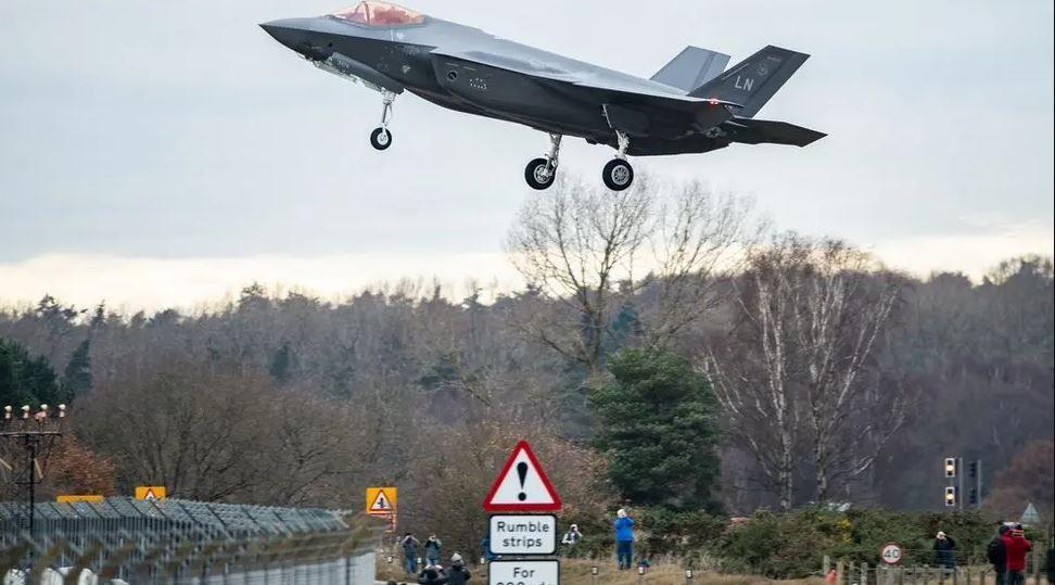 A United States fighter aircraft with a twin tail coming in to land at an RAF base. The perimeter fence can been seen as well as civilian aircraft spotters. There is a road sign in the foreground