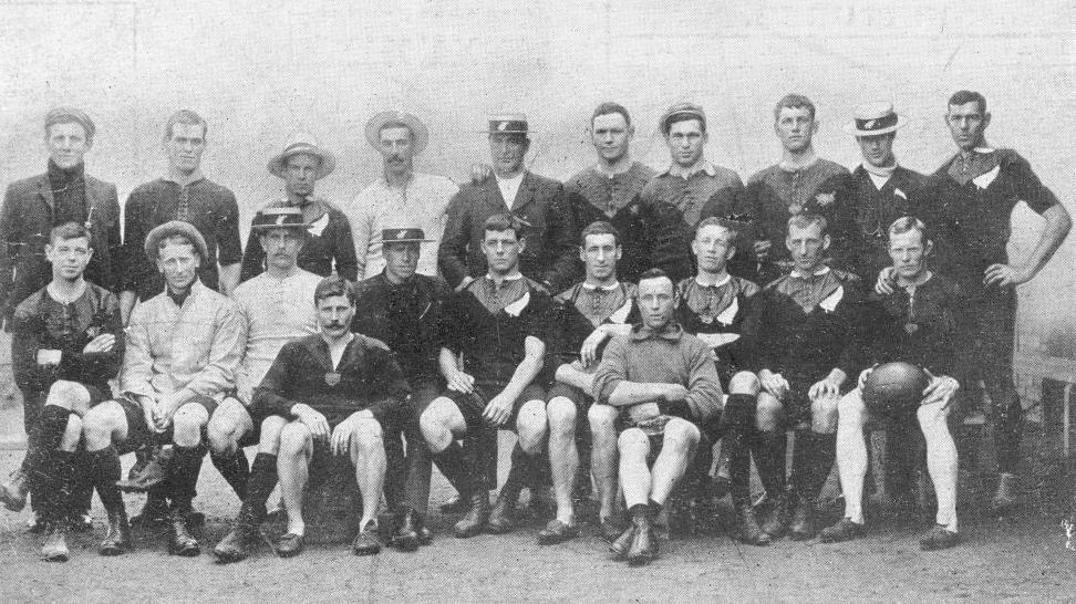A black and white picture of the All Blacks 1905 team - there is a group of men in black or white rugby tops, shorts and boots. 