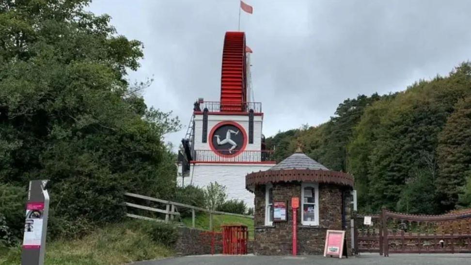 The front of the Laxey Wheel, which is a large white building with the red wheel mounted on it, which has a small round kiosk and a turnstile in front of it.