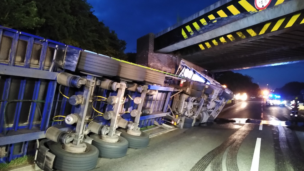 A large lorry is on its side after hitting the A5 bridge in Hinckley, because it's too big to get through. It's dark and you can see vehicle lights in the distance as other drivers can't get through due to the accident. 