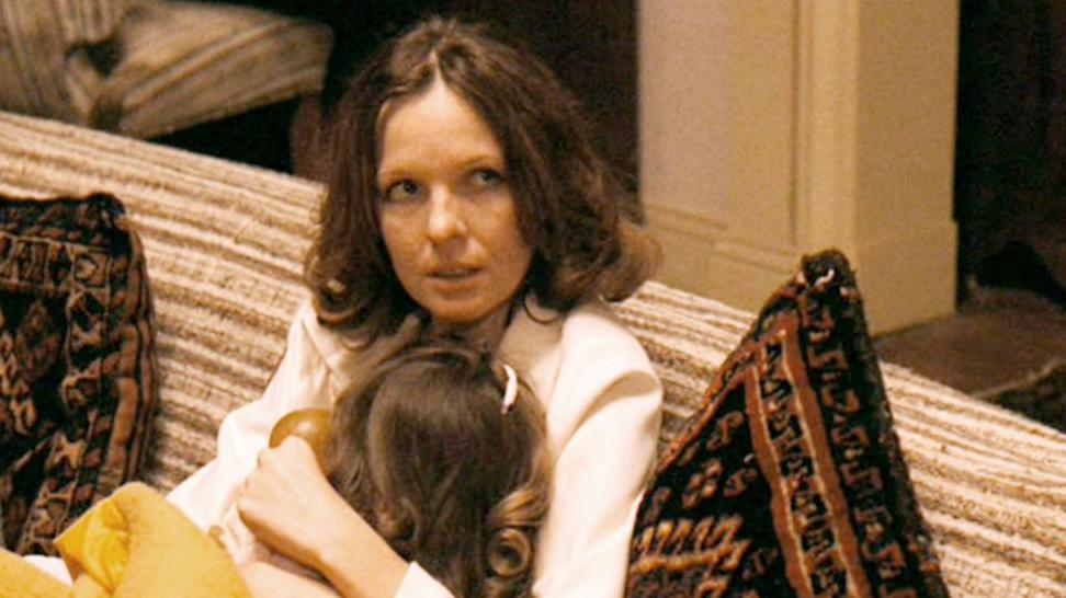 Diane Keaton sitting on a sofa in The Godfather Part II film, wearing a white long-sleeve shirt, holding a small girl against her chest.