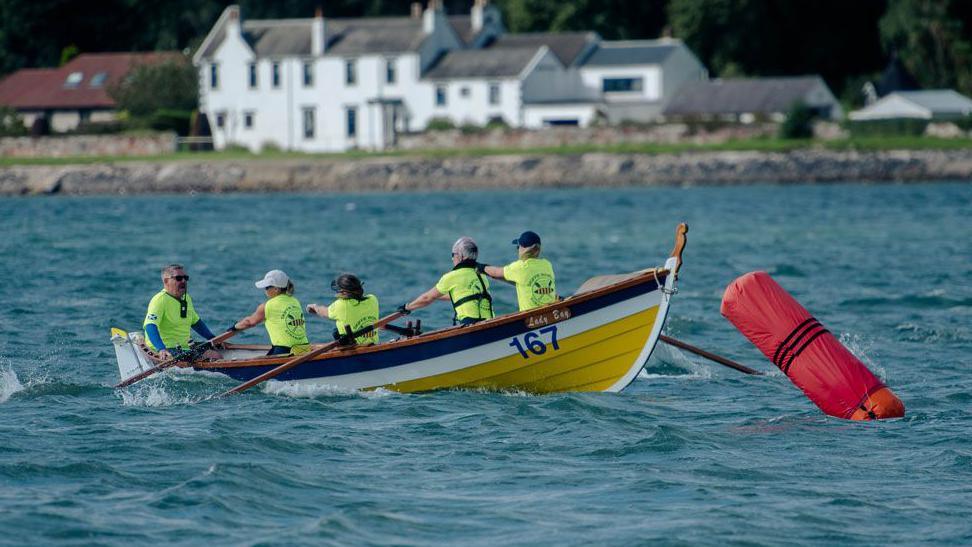 A crew in yellow tops ina a yellow, white and blue boat rounds a buoy in slightly choppy waters