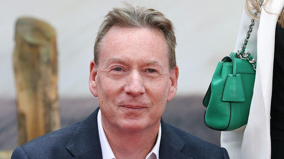 Frank Gardner. A man with blonde hair wearing a suit looking at the camera. There is a green hand bag to his left.