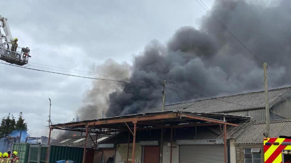 A large plume of smoke coming from an industrial unit in North Benfleet. Firefighters and a fire engine are at the scene. The roof is a metal structure. Firefighters are on an aerial ladder.