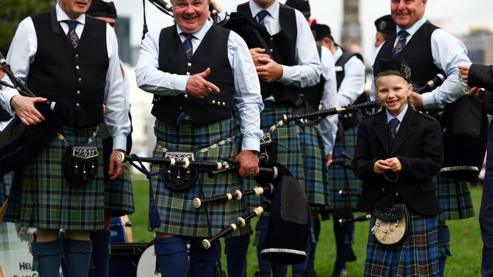 A group of pipers wearing kilts standing next to young girls smiling as she also wears a kilt.