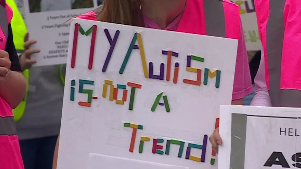 A person amidst a protest is holding a sign saying "My Autism is not a Trend!"