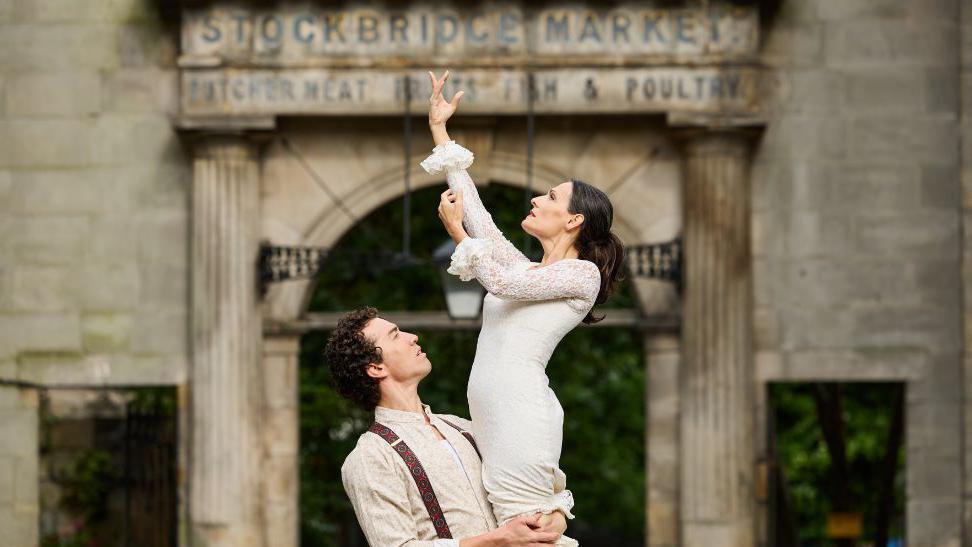 A man and a woman in traditional flamenco dancing clothes. The woman is wearing a white lace dress and is being lifted by the man. She has her arm raised in the air. The man is dressed in light beige clothing with suspenders. The background features a stone archway with the words "Stockbridge Market" above it