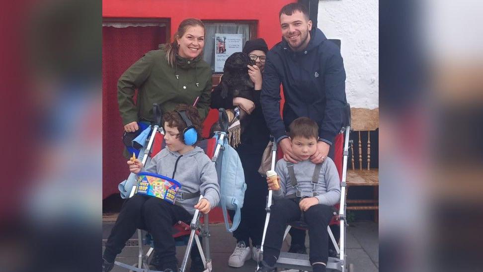 Heather Smith and her husband are photographed standing behind two of their sons, who are sitting in buggies. Their eldest son stands between them, holding a small black dog. 