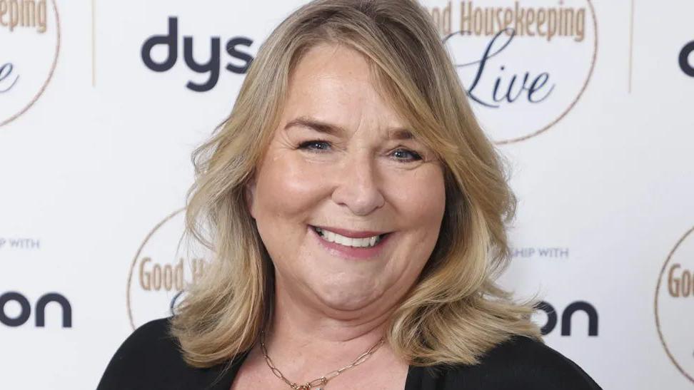 A head and shoulder shot of Fern Britton who has shoulder-length blonde hair and is wearing a black top and a gold necklace