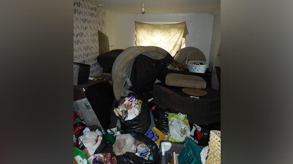 Room in flat shows light bulb with no lamp shade, bags of rubbish all over the floor and furniture in a pile, with a dirty white sheet covering the window 