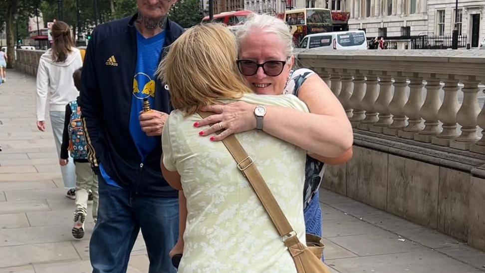Two women hugging on a London pavement, with a stone barrier behind them. other pedestrians are walking in the background and there are buses and a van on the road. The woman whose face can be seen is smiling over the shoulder of the other, who has her back to the camera.