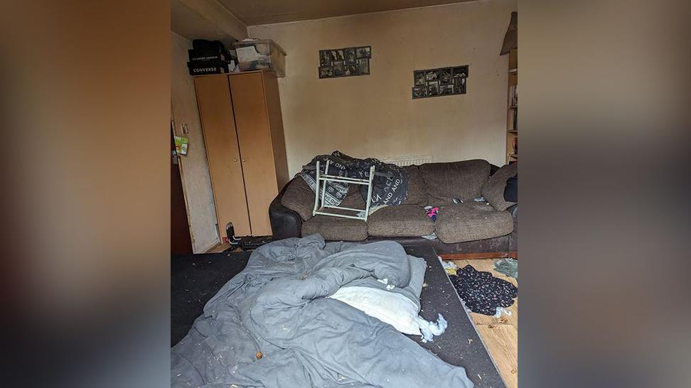 A messy house showing mess and rubbish with a dirty duvet on the floor near a brown sofa.