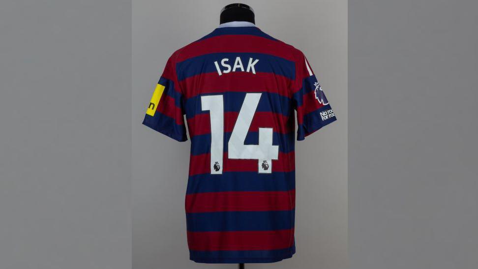 Dark red and dark blue striped Newcastle United shirt which says Isak and the number 14. It is on a mannequin.