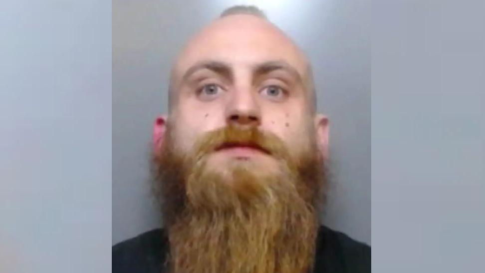 Mugshot of a man with a little hair in the middle of his head and a long brown beard staring at the camera. He is wearing a black shirt.