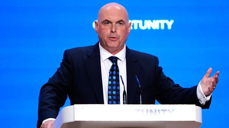 Paul Davies standing at a lectern in a navy suit, white shirt and navy tie with blue dots. He has a shaved head and is gesticulating as he talks during a conference. The backdrop is blue and the word opportunity is written on the lectern and projected onto the wall behind him.