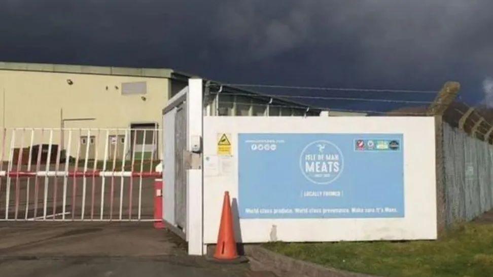 The exterior of Isle of Man Meats. It features a white high gate next to white  board with a blue sign that says Isle of Man Meats.