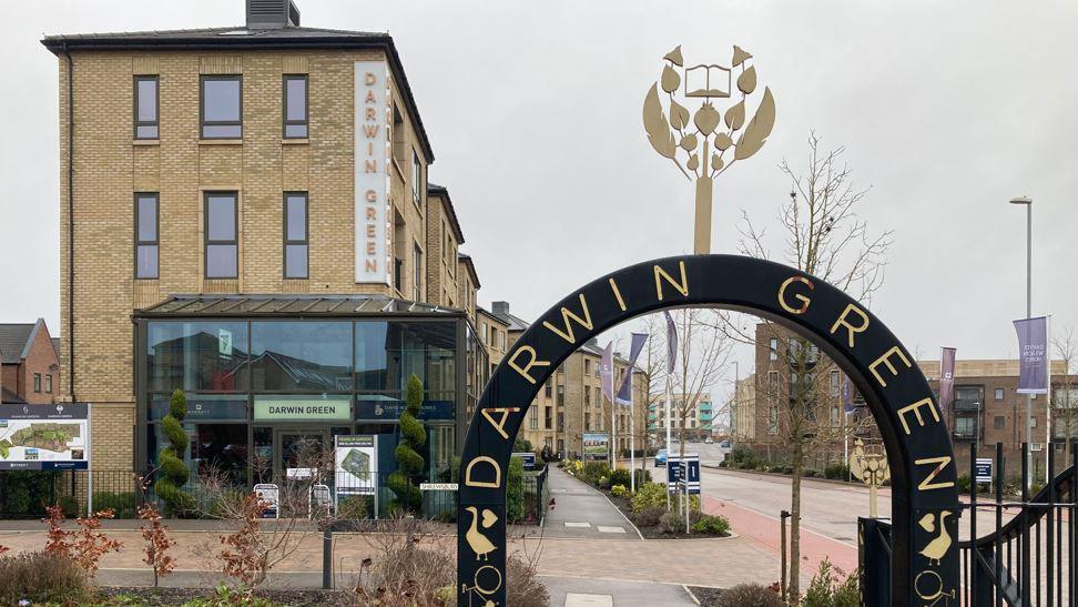 To the left is a 4 storey building with a glass front covering the first 2 floors. To the right is a black crescent shaped gate with Darwin Green written on it. Newly built homes, flags and bare trees are behind it.   