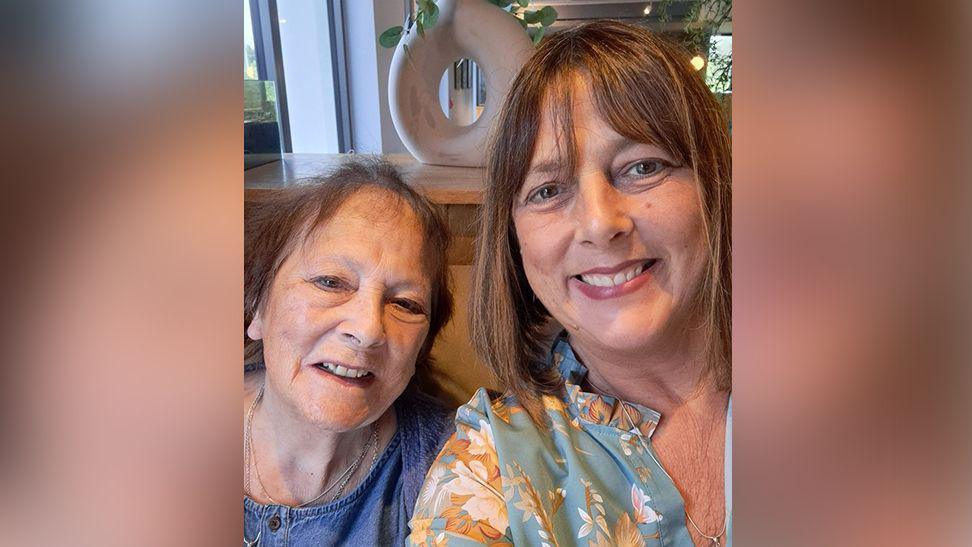 Christine Edwards (left) with brown hair and wearing a denim top with her daughter Amanda with shoulder length brown hair wearing a blue and flowered pattern blouse. They are both smiling.
