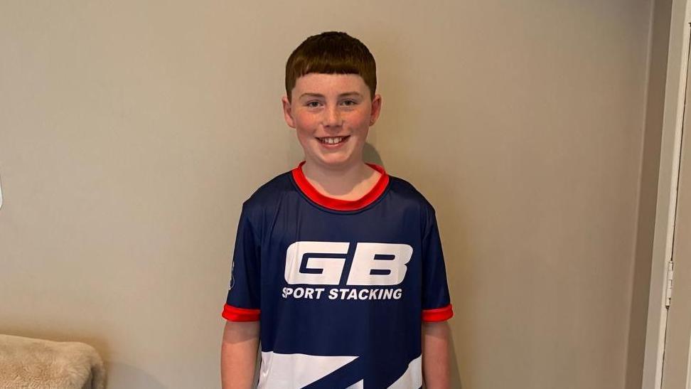Olly is a boy with brown hair and is wearing a blue sports top with "GB sport stacking" written on it in white text. He is standing in front of a wall in a living in front of a cream-coloured wall.