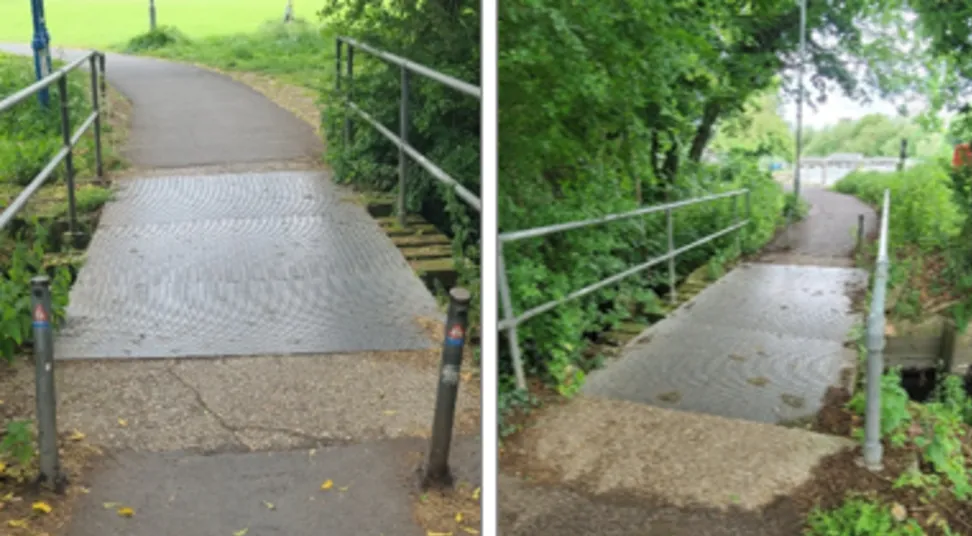 The old bridge, which is clearly worn and old, with planks of concrete above a metal section, with metal railings across it with foliage and other trees behind it on the left side of the bridge 