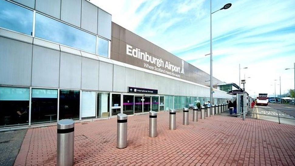 The outside of Edinburgh Airport. Metal bollards protecting the airport run along the foreground. in the background is the Edinburgh Airport sign and the entrance doors.