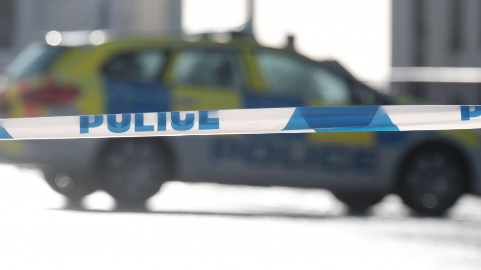 A police cordon tape with blue writing with a police car out of focus in the background.