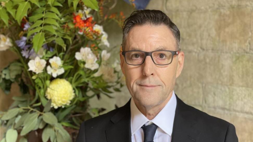 Daryl Slater in a black suit and tie with his hair slicked back and glasses on standing in front of a flower display