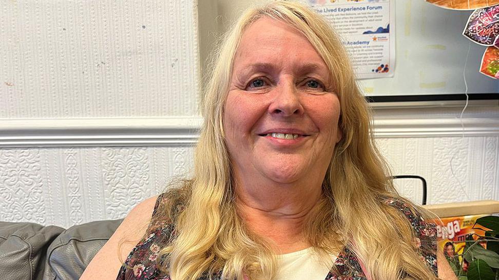 Debra Etherington is sitting on a grey leather-look sofa in front of a white wallpapered wall. She has long, blonde hair and is wearing a sleeveless dark purple patterned top or dress. Just behind her, hardly visible, is a box for the block-stacking game Jenga.