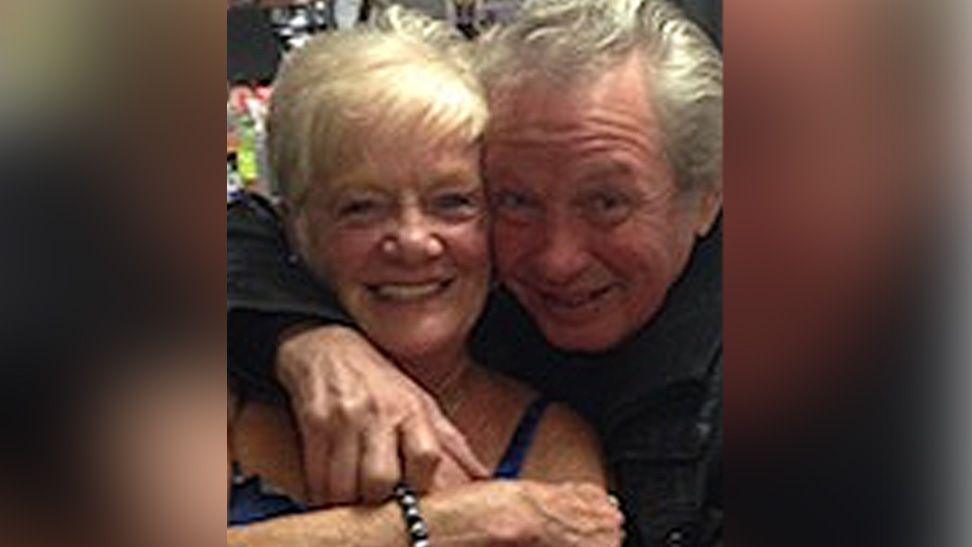 Sheila Jackson with blond hair smiles as Eric Greener with grey hair has his arm around her neck in front of a bar. Eric is also smiling.