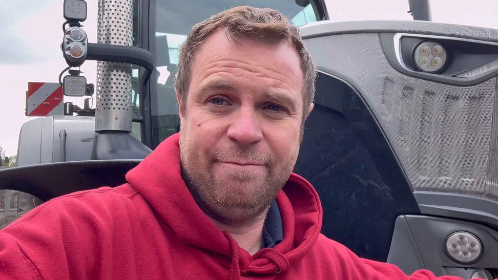 Olly Harrison has short hair and a short beard. He is wearing a red hooded top and is stood in front of a grey tractor.