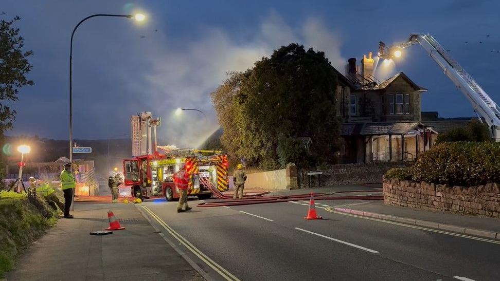 Large derelict hotel on the corner of two roads - several fire engines are to the side with numerous firefighters
