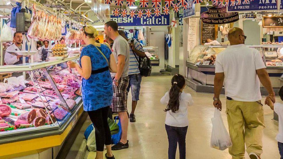 People in a market hall indoors. Several people are in between market stalls including a butchers with meat on display. A woman and a man look at the meat while a man and a girl walk away from the camera, the man carrying a bag in his left hand.