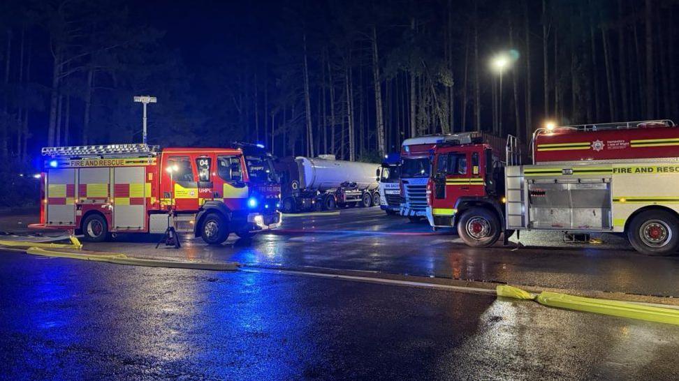 Two fire trucks parked on tarmac with a tanker in the background. It is night time