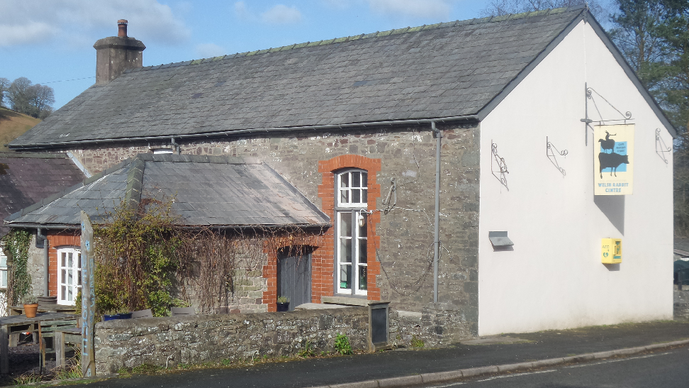 The outside of the Welsh Rarebit Centre. It is an old renovated school and on the right is a sign saying the Welsh Rarebit Centre. The picture has been taken on a sunny day.