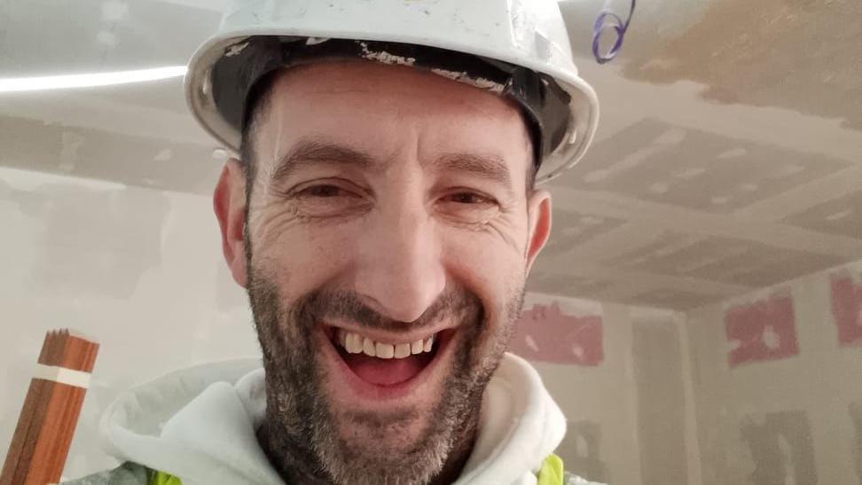 Andrew smiling at the camera. He is wearing a white hard hat, a white hoodie and a high-vis jacket. He is standing in a room with walls that have yet to be decorated.