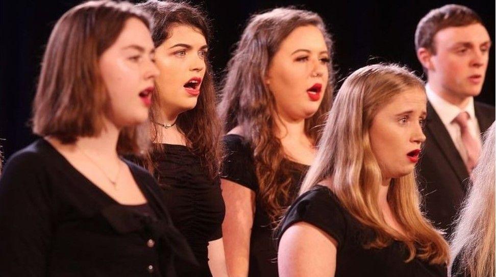 Shows four young woman and a young man - dressed in black - singing