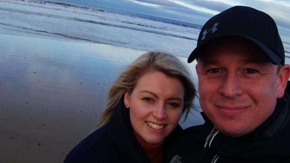 A selfie of Samantha and Trevor. They're standing on a beach and smiling into the camera. Samantha has her hair down and Trevor is wearing a black cap.