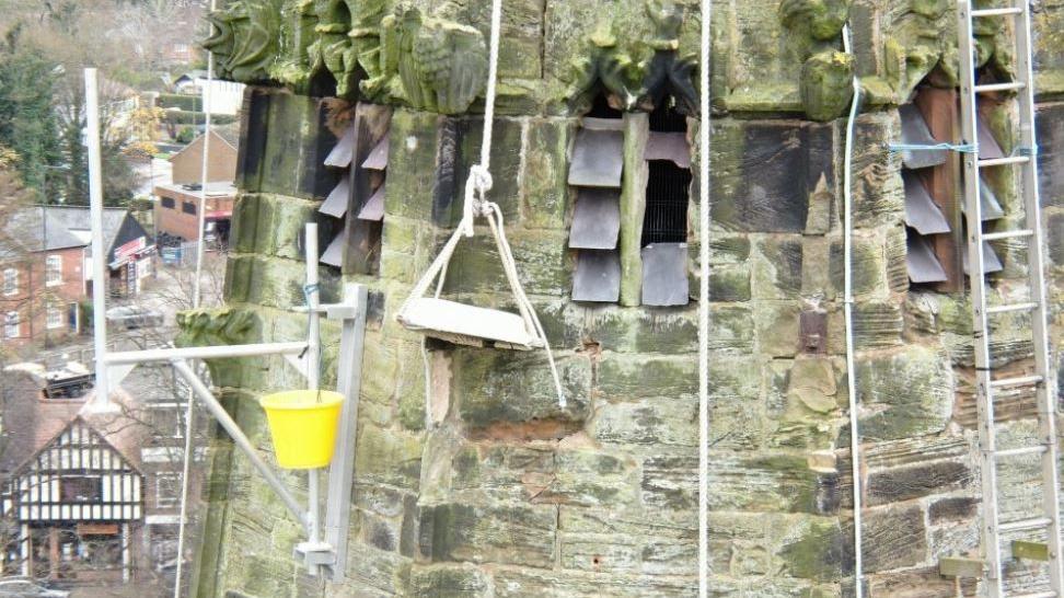 A wooden chair suspended by ropes with a yellow bucket also suspended nearby, both hanging from the side of a stone church steeple with small buildings visible below