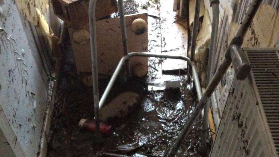 The entryway of a home with thicj mud on the floor and up the walls, with various brooms and mops in the middle and a radiator on one side