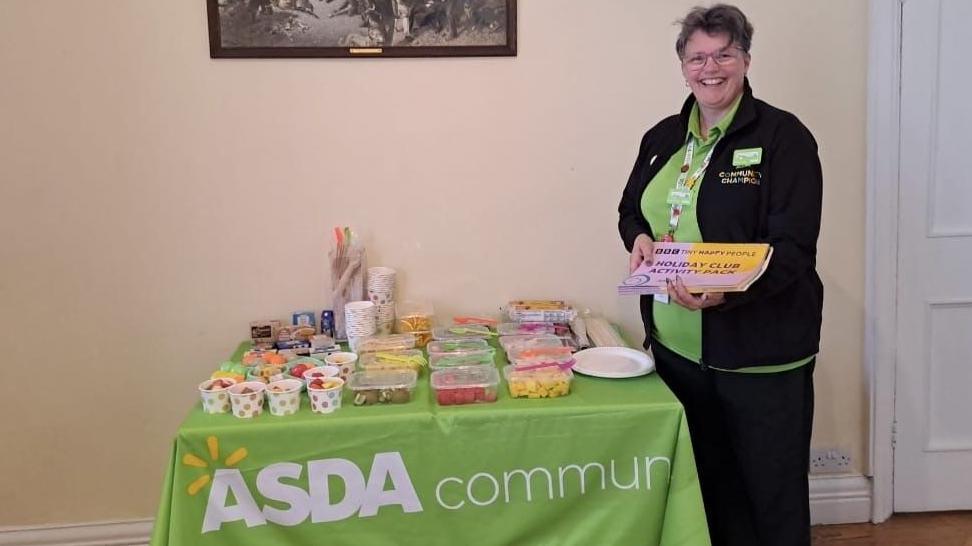 Donna Robinson wearing her Asda black fleece and green top uniform. She is standing next to a table which has cakes and sweets on, and reads 'Asda community'. She is holding a pink and yellow holiday club activity pack in her hands.