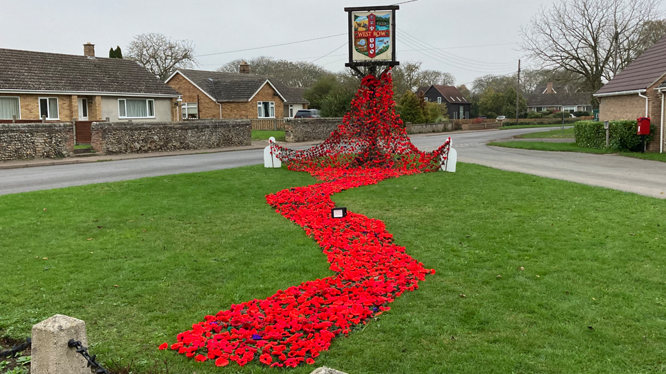 The village green in West Row, surrounded by bungalows, with red knitted and crocheted poppies attached to camouflage netting forming a cascade from bottom of the village sign, across the grass and towards the camera