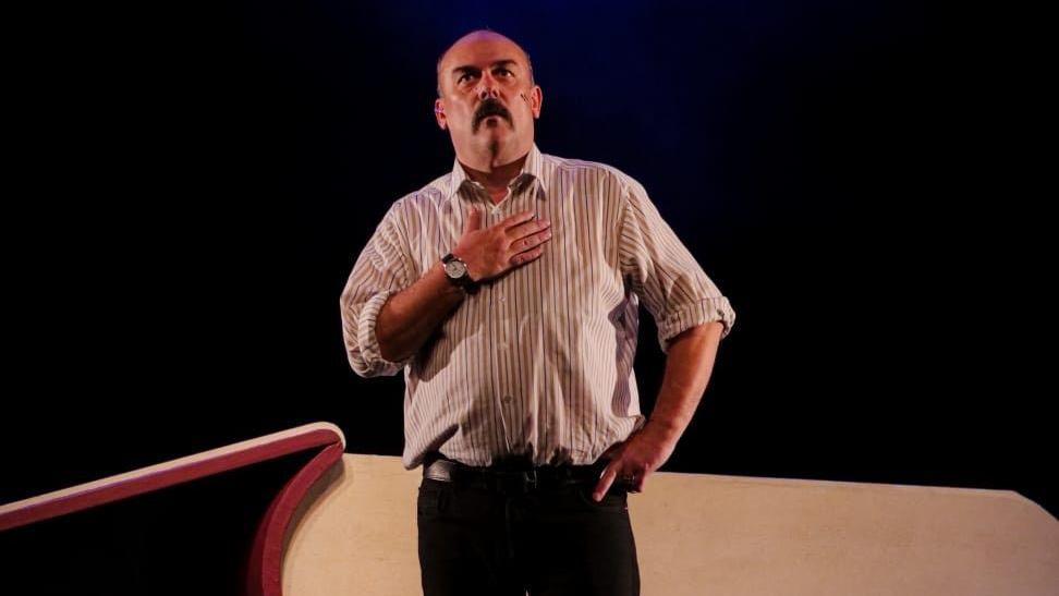 Paul Garrett on stage, with a moustache and brown hair wearing a white striped shirt and a watch