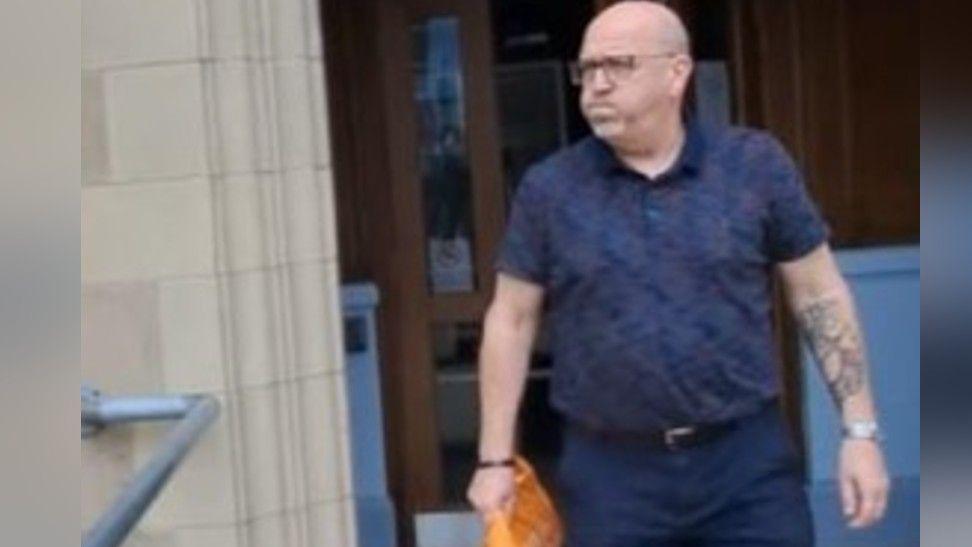 Mark Hodgson leaving court. He is wearing a blue short-sleeve shirt and blue trousers. He is bold, is wearing glasses and has a large tattoo on his left arm.