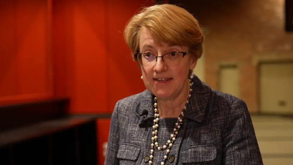 Woman with medium length red hair and glasses dressed in a smart jacket looks at camera